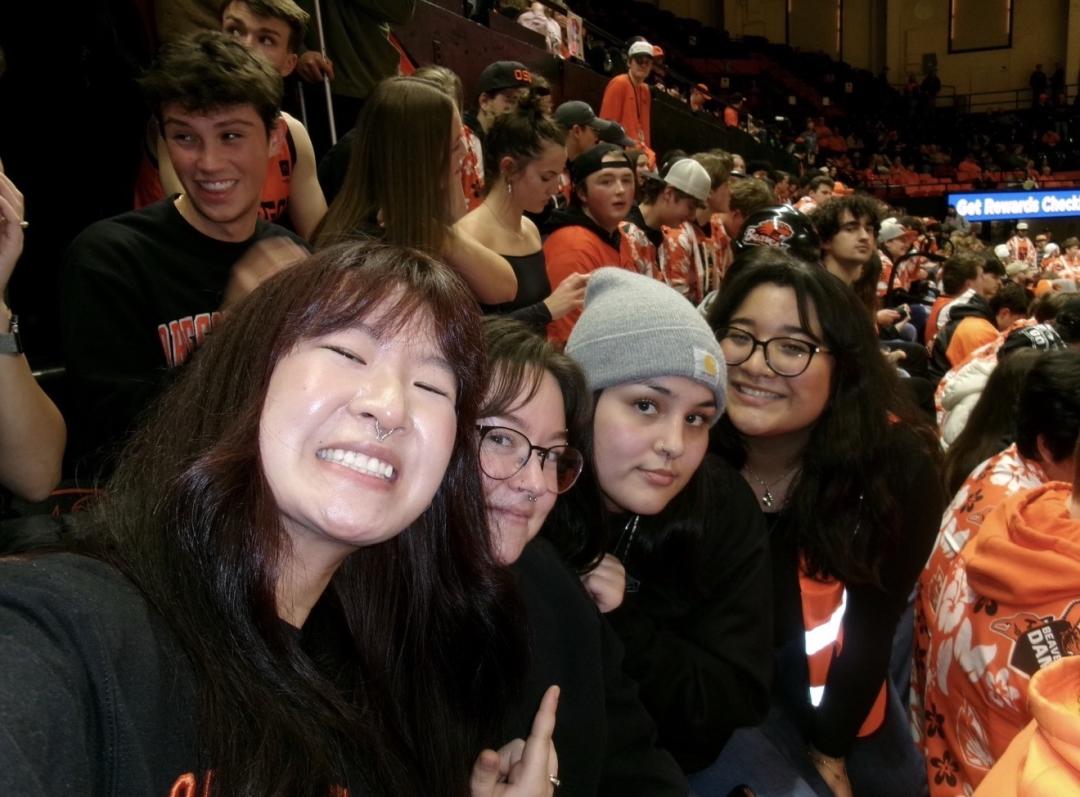 Four women smile for a picture in stadium stands packed with Beaver fans during a basketball game.