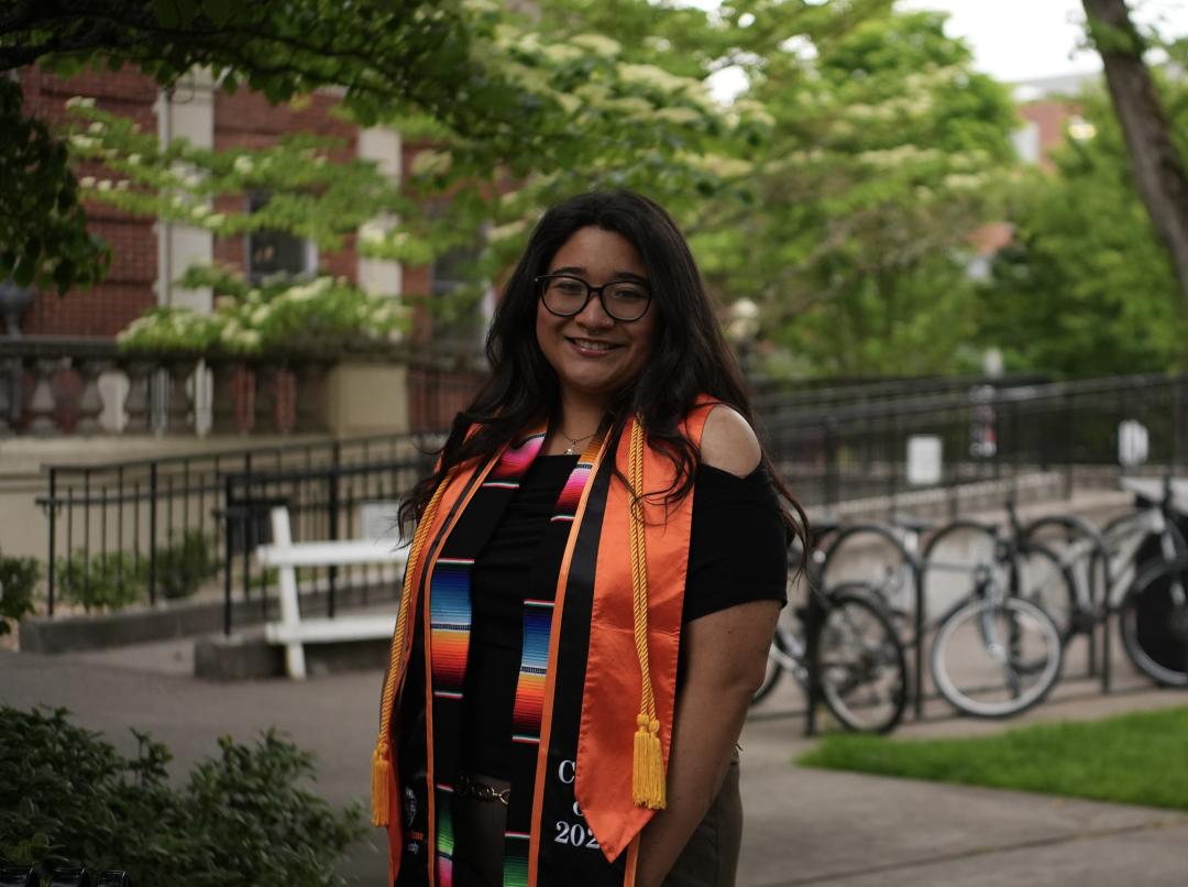 A woman with glasses smiles in a black dress with OSU and cultural graduation regalia amidst a sunny campus backdrop.