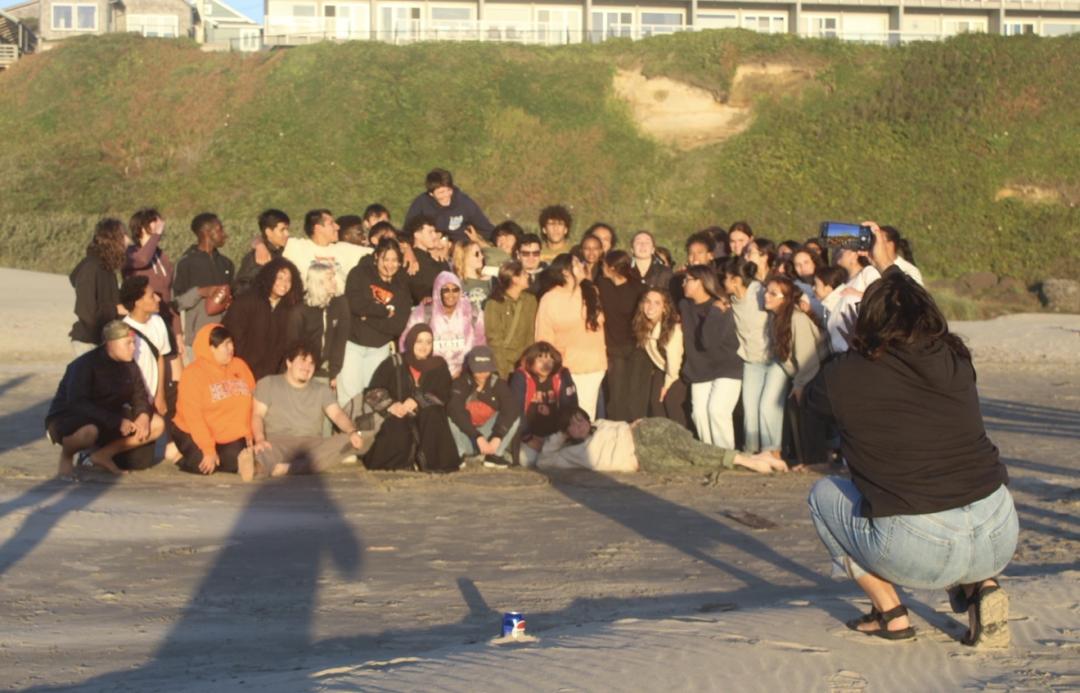 Under a setting sun, a large group of students huddle on a beach while a woman in the foreground kneels to take their picture.