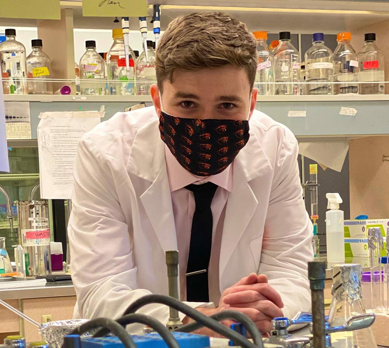 A young man in a lab coat and face mask leaning over a counter with test tubs