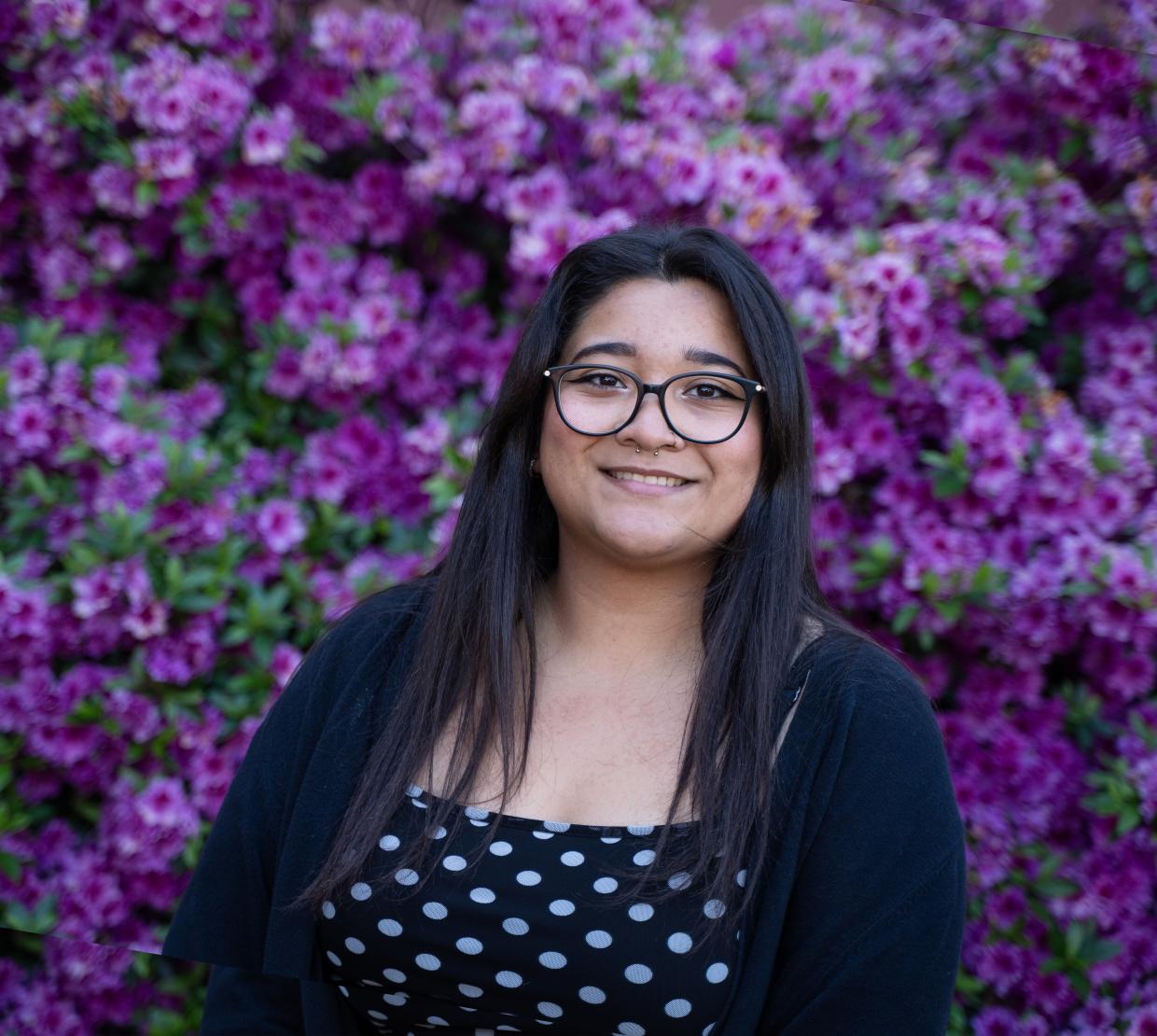 A dark-haired woman smiles in front of violet flowers, wearing round glasses, a black-and-white polka dot shirt, and two small piercing on her nose.