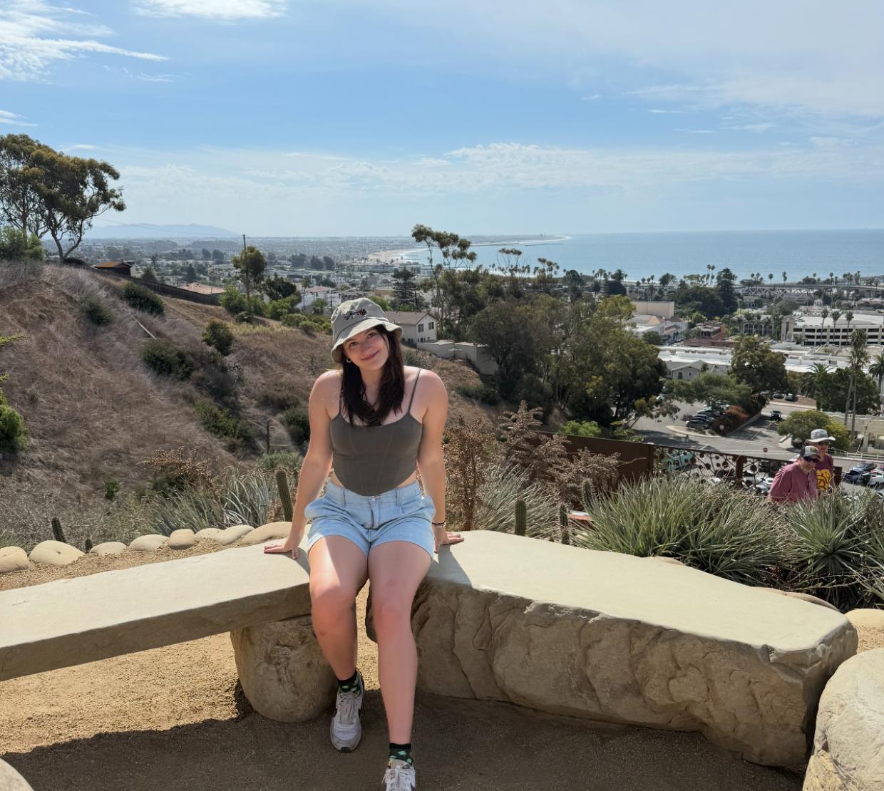 A woman in shorts and a hat poses for a photo near the ocean.