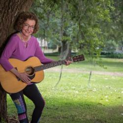 A woman in a purple shirt leaning against a tree and holding a guitar