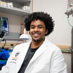 A man wears a lab coat with the OSU crest, sitting in a lab beside a microscope and various equipment.