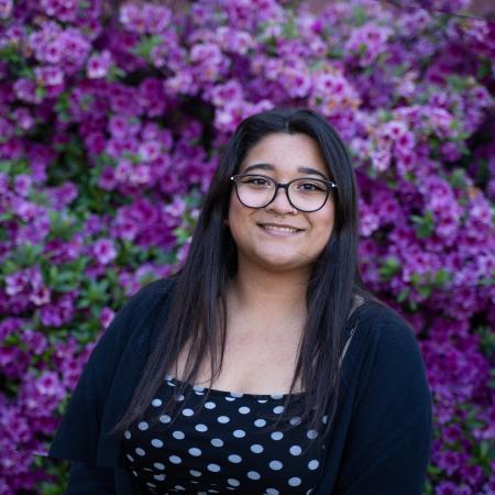 A dark-haired woman smiles in front of violet flowers, wearing round glasses, a black-and-white polka dot shirt, and two small piercing on her nose.