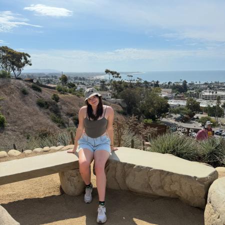 A woman in shorts and a hat poses for a photo near the ocean.