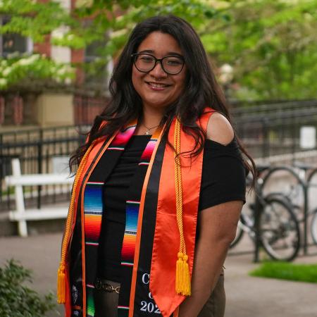 A woman in glasses and graduation regalia smiles brightly in front of a sunny campus backdrop.