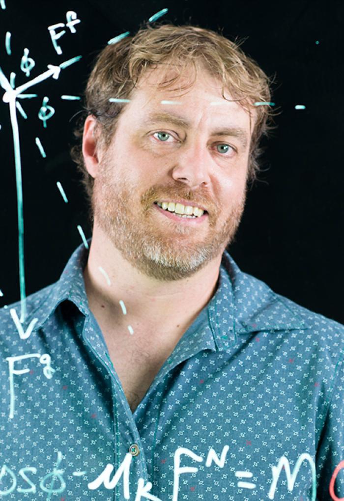 Kenneth C. Walsh smiles in a headshot behind a a clear whiteboard with physics equations.