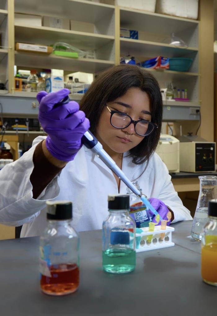 A woman wearing glasses and a white lab coat focuses on pipetting brightly colored solutions into small tubes in the lab.