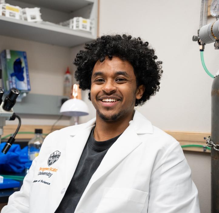A man wears a lab coat with the OSU crest, sitting in a lab beside a microscope and various equipment.