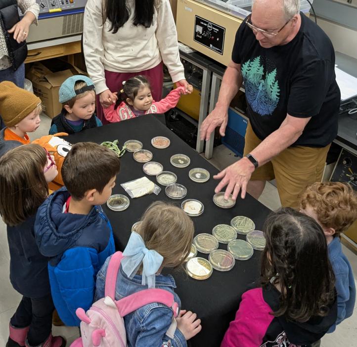 A male scientist gestures to fungi growng in petri dishes as preschoolers look on