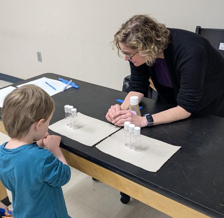 A woman scientist leans in to speak to a preschooler as they look at jars with flies