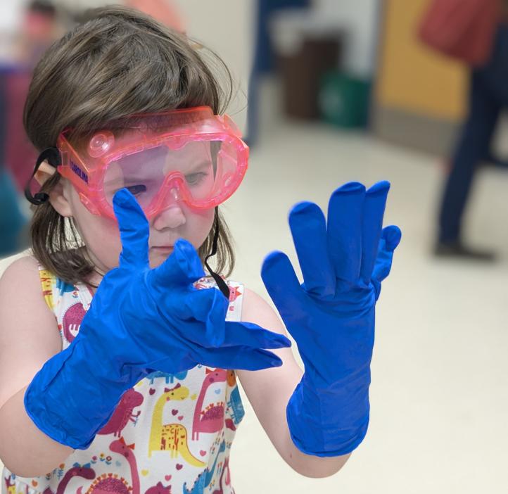 A young child in bright safety goggles considers the too-large safety gloves on her hands